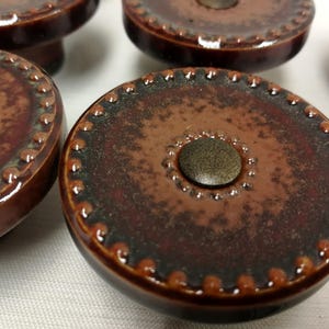 Może przedstawiać: Zestaw okrągłych ceramicznych gałek do szafek z błyszczącym brązowym szkliwem i nakrapianą fakturą. Każda gałka posiada wypukły koralikowy brzeg wzdłuż zewnętrznej krawędzi oraz mniejszy koralikowy okrąg wokół centralnego metalicznego detalu.