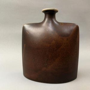 以下が含まれることがあります： 丸みを帯びた四角い形をした、濃い茶色の陶器の花瓶。首の部分は少し広がっています。つや消しの仕上げで、細かい斑点模様があります。