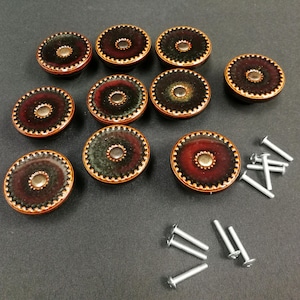 May include: A set of nine circular cabinet knobs featuring a gradient of deep red, black, and green tones with a metallic copper-colored scalloped edge and a central silver-toned stud, accompanied by nine silver-toned metal mounting screws.