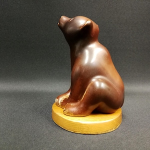 Pode incluir: Uma figura de urso de cerâmica marrom sentada em uma base redonda de madeira clara. O urso está sentado, olhando para cima. A figura tem um acabamento brilhante e está em um fundo escuro.