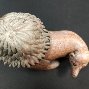 Pode incluir: Estatueta de cerâmica de um ouriço com um dorso texturizado e detalhado e um corpo castanho-rosado. O ouriço está enroscado, com a cabeça e o focinho apontando para baixo. A estatueta tem um aspeto vintage.