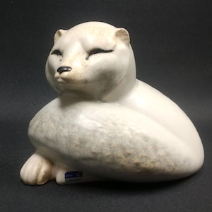 May include: A white ceramic sculpture of a curled up arctic fox. The sculpture has a textured, speckled finish and a blue and white sticker on the bottom.