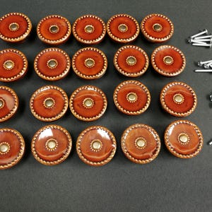 May include: A set of twenty round, glossy, burnt-orange ceramic cabinet knobs with a textured, beaded edge and a central metallic gold-toned circular inlay. The set includes twenty matching silver-toned metal mounting screws for installation.