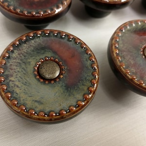 May include: A set of round ceramic cabinet knobs featuring a glossy glaze in shades of deep red, brown, and muted green. Each knob has a raised beaded border along the outer edge and a central circular metal accent with a textured bronze finish.