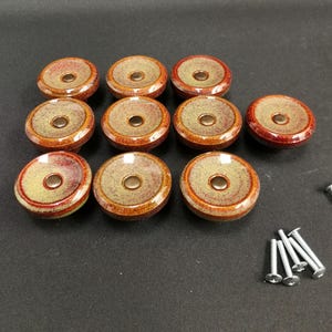 Op de afbeelding: Een set van tien ronde keramische kastknoppen met een gespikkeld beige midden en een diepe roodbruine geglazuurde rand. Elke knop heeft een centraal metalen schroefgat. Tien zilverkleurige metalen montageschroeven zijn inbegrepen.