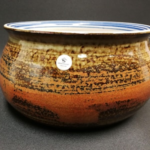 May include: A brown ceramic bowl with a blue and white striped rim. The bowl has a textured, layered glaze with brown, orange, and black tones.