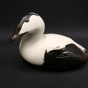 May include: A white and black hand-carved wooden duck decoy. The duck has a black head and a white body with black markings. The decoy is a popular item for bird watchers and collectors.