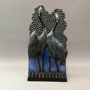 May include: A ceramic art piece featuring two stylized cranes with long necks and beaks, in shades of gray and black. The birds are set against a textured background of small, dark blue and green dots, with a blue base.