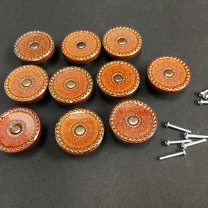 May include: A set of nine round cabinet knobs with a speckled orange and brown glazed finish, featuring a central metallic stud and a textured beaded border. The set includes nine silver-toned metal mounting screws for installation purposes.