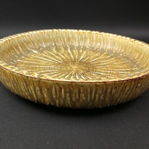May include: A round ceramic bowl with a textured, radial pattern in shades of tan and brown. The bowl's rim has a darker brown edge, and the interior features a sunburst design. The bowl is suitable for serving food.