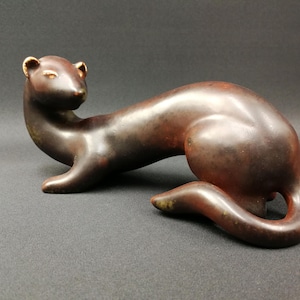 Könnte beinhalten: Eine Bronzeskulptur eines schlanken Tieres, möglicherweise ein Wiesel oder Frettchen, in liegender Pose. Die Skulptur hat eine glatte, polierte Oberfläche mit einer satten, dunkelbraunen Farbe und subtilen goldenen Akzenten an den Ohren. Die Form des Tieres ist länglich und anmutig.