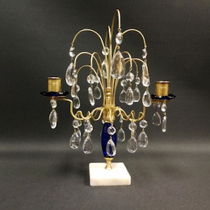 May include: A two-arm brass candelabra featuring a central blue glass column, a white square marble base, and numerous clear teardrop-shaped crystal prisms hanging from curved brass arms. The candle holders are accented with dark blue glass bobeches.
