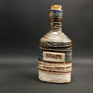 Op de afbeelding: Een handgemaakte keramische fles met een kurk, voorzien van een getextureerd oppervlak met horizontale ribbels in tinten bruin, blauw en wit. Een rechthoekig label aan de voorzijde is voorzien van het woord SNAPS in donkere hoofdletters.