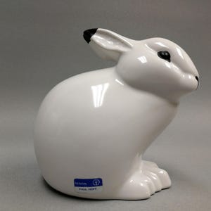 May include: A white ceramic rabbit figurine with black accents on the ears and eyes. The glossy finish reflects light, highlighting the smooth curves of the sculpture. A blue and white sticker with text is on the base. A decorative item.
