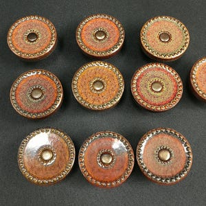 May include: A set of nine circular ceramic buttons featuring a mottled orange, brown, and red glaze. Each button has a raised central circular detail and a textured, beaded border around the outer edge, creating a consistent, uniform appearance.