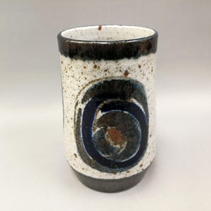 May include: A cylindrical ceramic mug with a speckled white exterior, a dark brown rim and base, and a central spiral design in shades of blue, brown, and gray. The mug is approximately 5 inches tall.
