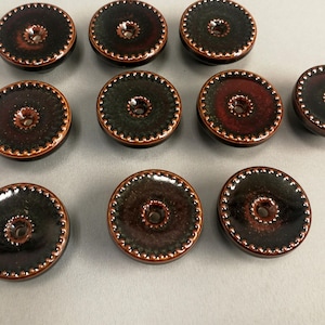 Op de afbeelding: Een verzameling van tien keramische knopen. De knopen zijn rond met een donkerbruin midden en een decoratieve rand. De randen zijn lichtbruin van kleur. Elke knoop heeft een gat in het midden om te naaien.