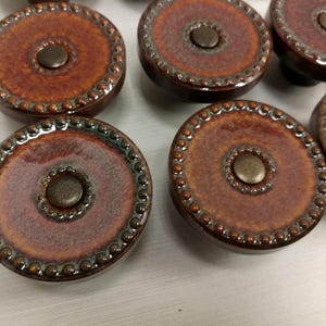 May include: A collection of round ceramic cabinet knobs featuring a glossy, mottled brown and rust-colored glaze. Each knob has a raised, beaded texture along the outer edge and a central circular indentation containing a small, dark metal stud.