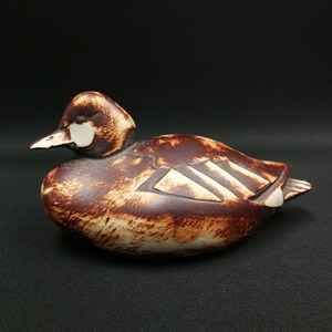 May include: A ceramic duck figurine in shades of brown and cream. The duck has a detailed design with a white beak and accents. The figurine is resting on a dark surface, showcasing its form and texture.