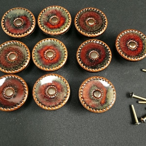 May include: A set of ten round cabinet knobs featuring a glossy, multi-colored glaze in shades of deep red, forest green, and black with a textured, beaded edge. Each knob has a central metallic button and includes a matching metal mounting screw.