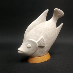 Könnte beinhalten: Weiße Keramikfigur eines Fisches mit strukturierter Oberfläche. Der Fisch steht auf einem Holzsockel.