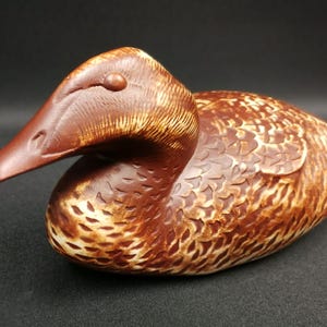May include: A hand-carved wooden duck decoy with brown and white paint. The duck has a detailed feathered pattern and a realistic beak.