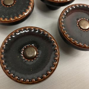 May include: A set of round cabinet knobs featuring a dark brown, glossy glazed finish. Each knob has a raised beaded border along the outer edge and a smaller beaded circle surrounding a central, textured, metallic-toned circular insert.