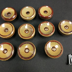 May include: A set of ten round ceramic cabinet knobs featuring a glossy tan center with a dark brown rim and a central metallic screw hole, accompanied by ten silver-colored metal mounting screws for installation on furniture or cabinetry.