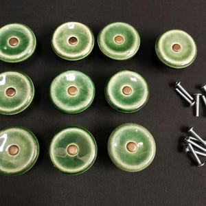 Könnte beinhalten: Ein Set aus elf runden Keramik-Schrankknöpfen mit glänzender grüner Glasur, einem helleren grünen Farbverlauf zur Mitte hin und einer kleinen metallisch braunen, kreisförmigen Einlage in der Mitte. Acht silberfarbene Metallschrauben liegen bei.