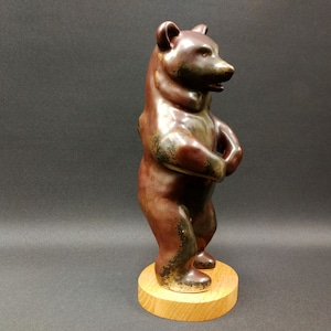 Op de afbeelding: Een bruin keramieken berenbeeldje dat rechtop staat op een ronde houten basis. De beer heeft een glanzende afwerking met donkere accenten en poseert met gevouwen poten. Het beeldje is een decoratief item.