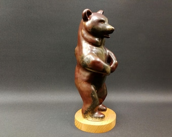 May include: A brown ceramic bear figurine standing upright on a round wooden base. The bear has a glossy finish with darker accents and is posed with its paws clasped. The figurine is a decorative item.