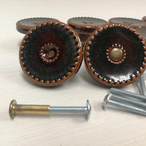 May include: A set of round cabinet knobs featuring a dark brown and black glazed finish with a textured, beaded edge. Each knob has a central brass-toned metal insert. Several silver-colored metal mounting screws are included with the hardware.