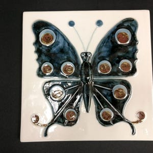 May include: A square ceramic tile featuring a butterfly design. The butterfly has dark blue wings with brown and white circular patterns. The tile is white with a glossy finish, and the butterfly's body is black.