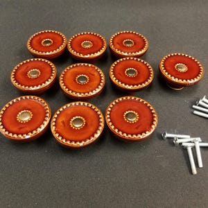 May include: A set of ten round cabinet knobs with a glossy burnt orange finish, featuring a central metallic disc and a beaded outer edge. The knobs are accompanied by ten silver-colored metal mounting screws for installation purposes.