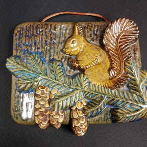 May include: A ceramic wall plaque depicting a squirrel on a branch with pine cones. The squirrel is brown and gold, set against a textured background. The leaves are green and blue, with brown pine cones. A red string is attached for hanging.
