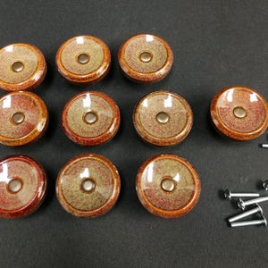 May include: Ten round ceramic cabinet knobs featuring a glossy finish with a speckled blend of reddish-brown and muted green tones. Each knob has a central metallic circular inlay and is accompanied by a set of matching silver-toned metal screws.