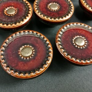 Può includere: Una collezione di pomelli rotondi per mobili con una finitura smaltata color bordeaux intenso e nero. Ogni pomello presenta un bordo esterno testurizzato a perline e un elemento centrale circolare liscio di tonalità metallica.