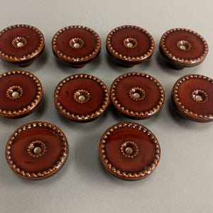 May include: Ten brown ceramic cabinet knobs with a circular design. Each knob features a central white dot surrounded by a ring of small, raised dots. The knobs are a warm, reddish-brown color, suitable for furniture restoration or DIY projects.