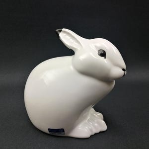 May include: A white ceramic figurine of a rabbit with black eyes and nose. The rabbit is sitting with its head tilted to the side.