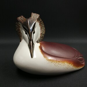 May include: A ceramic bird figurine with a white body, a brown back, and a detailed head. The head features a dark beak and a textured, feathery design. The bird is in a resting position, with a smooth, rounded form.