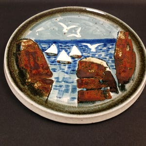 Op de afbeelding: Een rond keramieken bord met een handgeschilderd kustlandschap. Het ontwerp toont donkerbruine rotskliffen aan de zijkanten, een diepblauwe zee met witte zeilboten en een lichtblauwe lucht met witte vogels boven het wateroppervlak.