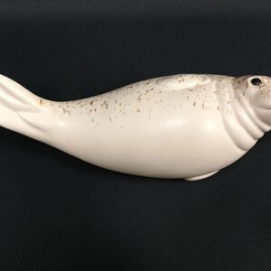 May include: A cream-colored ceramic seal figurine with brown speckles. The seal has a smooth, rounded body, a small head with black eyes, and a flipper-like tail. The figurine is lying on a black surface.