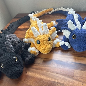 May include: Three handmade crochet dragons in black, yellow, and blue. The dragons have detailed features, including horns, wings, and large eyes. The yellow dragon has white accents, while the blue dragon has white accents. The black dragon has gray accents.