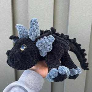 May include: A handmade black crochet dragon plush toy with gray accents on the horns, wings, and feet. The dragon has a large, golden eye and a textured, plush appearance. The toy is designed to resemble a fantasy creature.