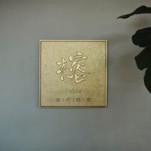 May include: A square, gold-colored sign with Chinese characters and the word "Table" in English. The sign has a textured surface and a simple, elegant frame. The sign is mounted on a gray wall.