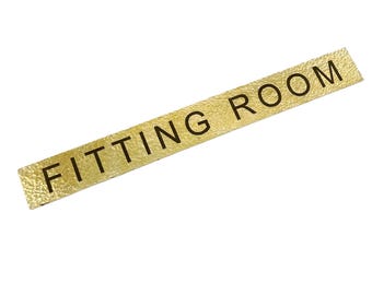 May include: A rectangular gold-colored sign with the words "FITTING ROOM" printed in black. The sign has a textured surface and is set against a white background. The sign is likely used in a retail setting.
