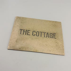 May include: A rectangular brass sign with the engraved text "THE COTTAGE". The sign has a textured surface and a slightly aged appearance. The lettering is in a simple, sans-serif font.