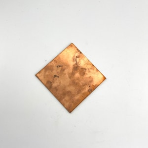 May include: A square copper sheet with a textured surface. The copper has a warm, reddish-brown colour with darker spots and variations in tone. The sheet is positioned diagonally against a white background.