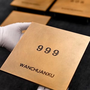 Könnte beinhalten: Ein quadratischer, goldfarbener Metallgegenstand mit der eingravierten Zahl "999" auf der Oberfläche. Der Text "WANCHUANXU" ist unter den Zahlen aufgedruckt. Der Gegenstand wird von einer behandschuhten Hand gehalten, mit anderen ähnlichen Objekten im Hintergrund.
