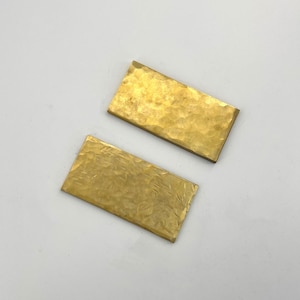 Può includere: Due pezzi rettangolari in ottone martellato con una superficie strutturata. L'ottone ha una calda tonalità dorata e i rettangoli sono posizionati su uno sfondo grigio chiaro. I pezzi sembrano fatti a mano.